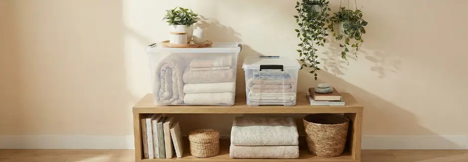 Pojemniki do przechowywania z poskładanymi tekstyliami na drewnianej półce, z roślinami i koszami.